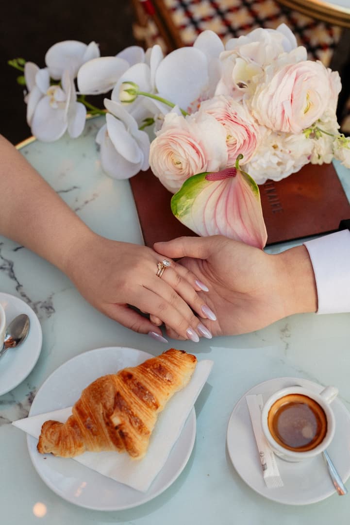 Their hands and wedding rings on a café table beside a croissant and an espresso