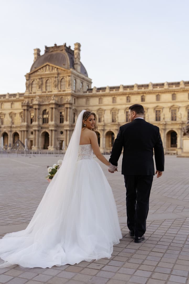 Aphrodite and Michael walking hand in hand across the Louvre courtyard at sunrise
