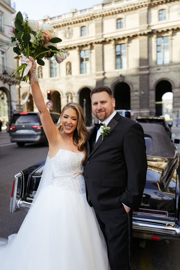 Aphrodite raising her bouquet beside Michael and the vintage car