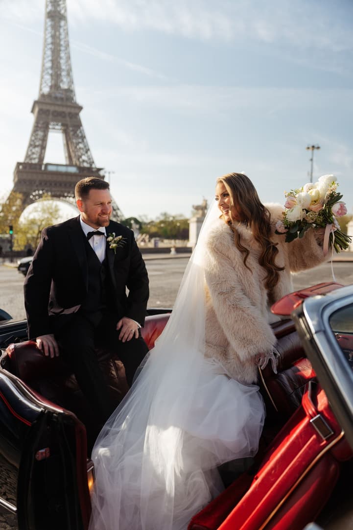 Aphrodite in a white fur stole in the vintage car with the Eiffel Tower behind
