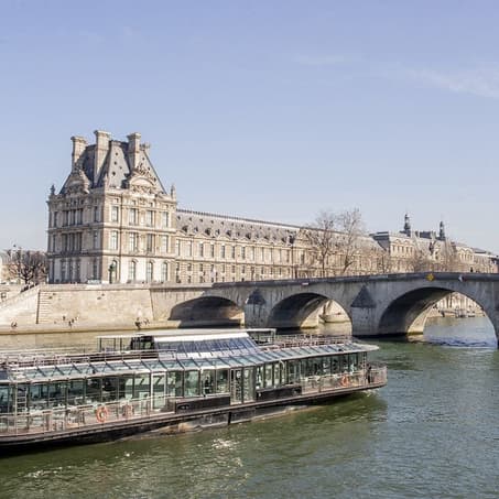The glass-walled boat of Ducasse sur Seine on the river, the Louvre behind it