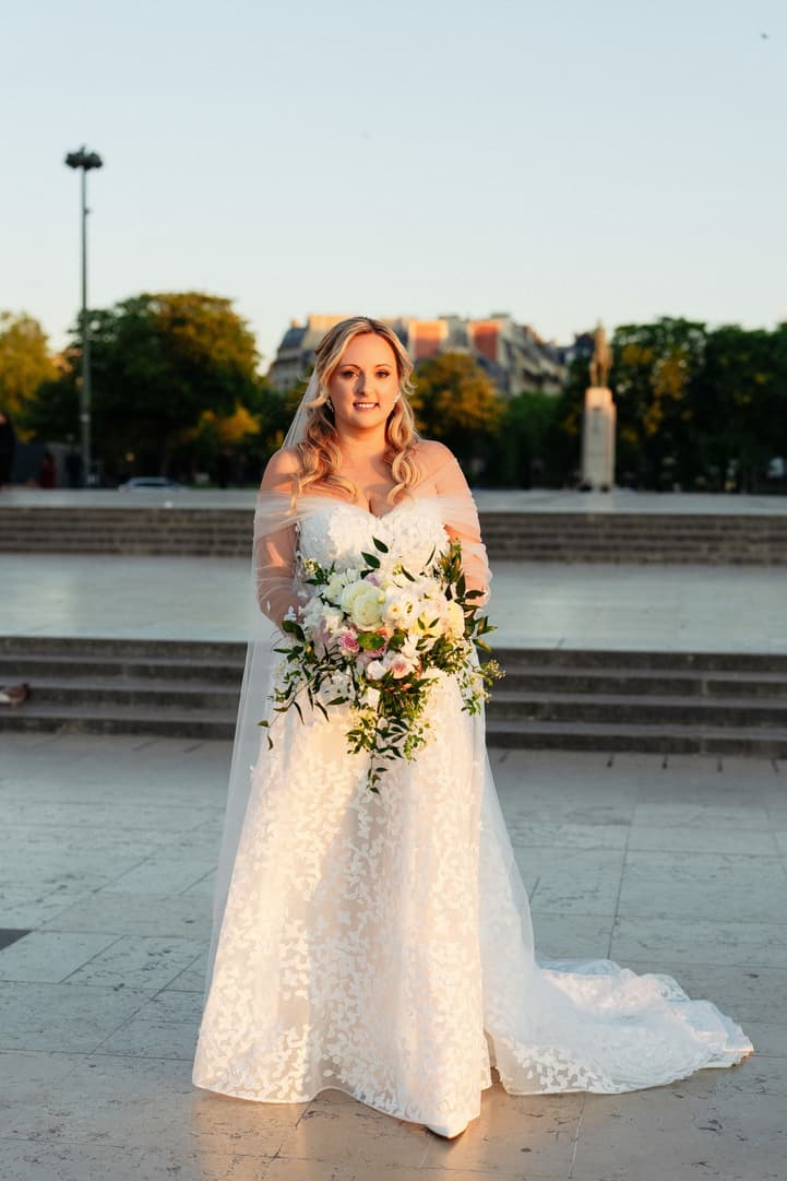 Victoria in her floral-sleeved wedding gown and veil, holding her cascading bouquet on the Trocadéro at sunrise