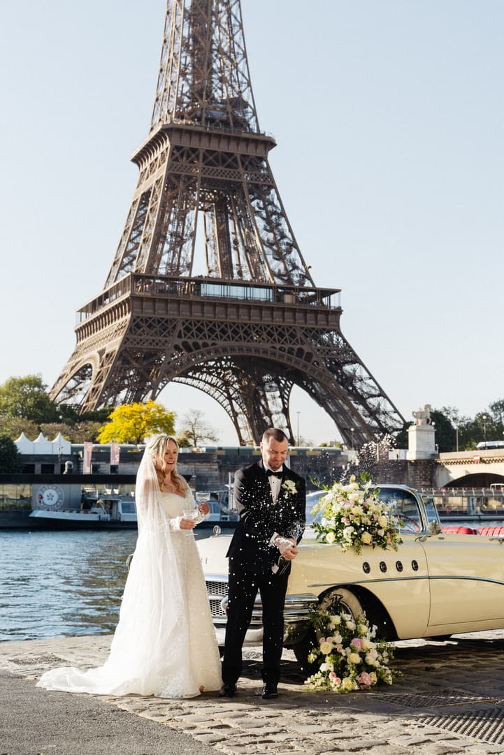 Victoria and Tyler open Champagne beside their white vintage car on the Seine, the Eiffel Tower behind