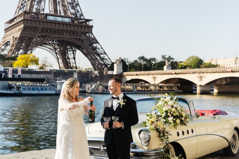 Victoria pours Champagne beside Tyler and the flower-dressed white vintage car on the bank of the Seine