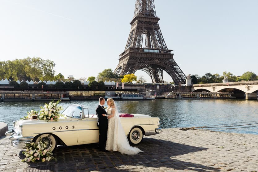 Victoria and Tyler by their white vintage car on the bank of the Seine, the Eiffel Tower behind