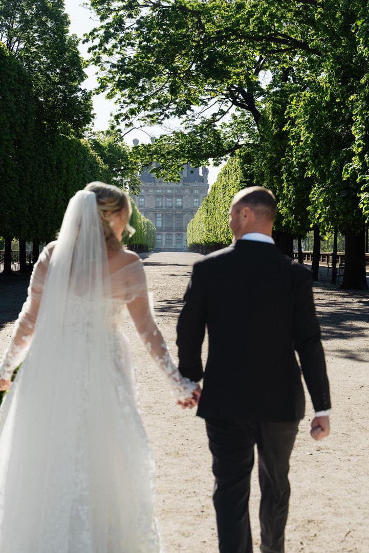 Victoria and Tyler walk hand in hand down a tree-lined allée of the Tuileries toward the Louvre