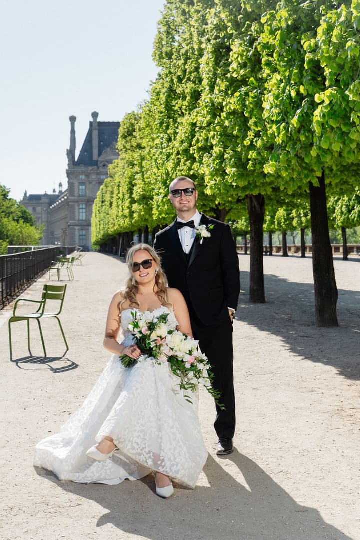 Victoria in sunglasses and her gown, Tyler behind her, by the green chairs of the Tuileries