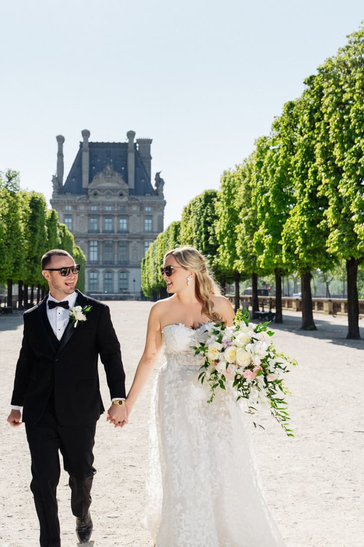 Victoria and Tyler in sunglasses walk hand in hand through the Tuileries, the Louvre behind