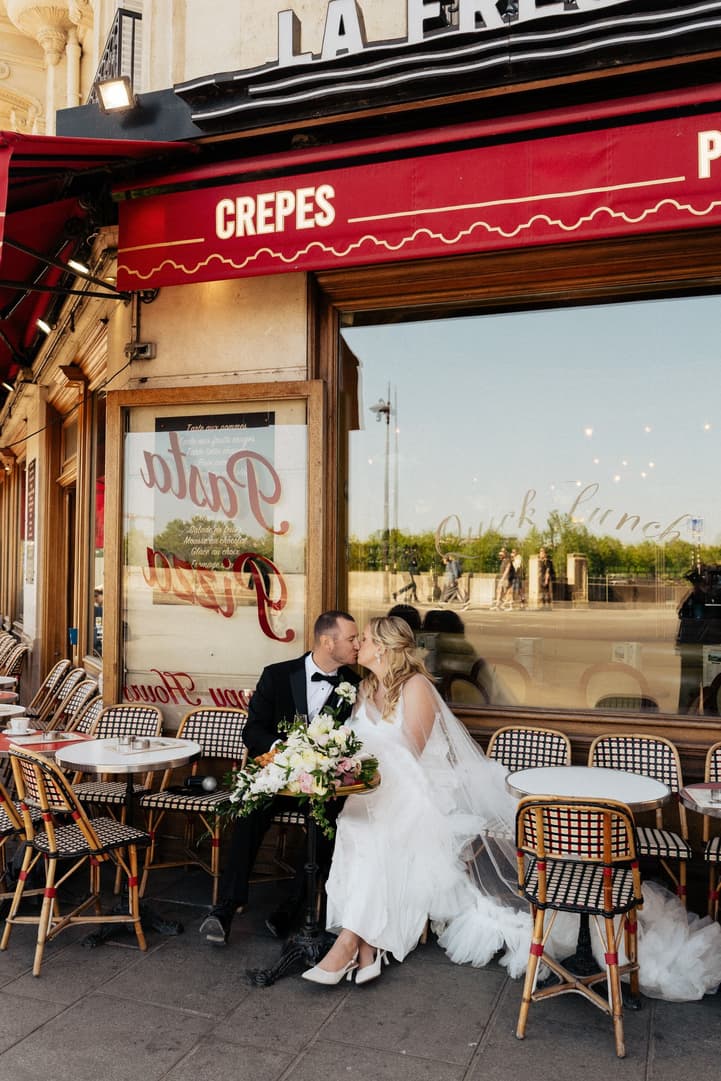 Victoria and Tyler kiss at a table on the terrace of La Frégate under its red awning