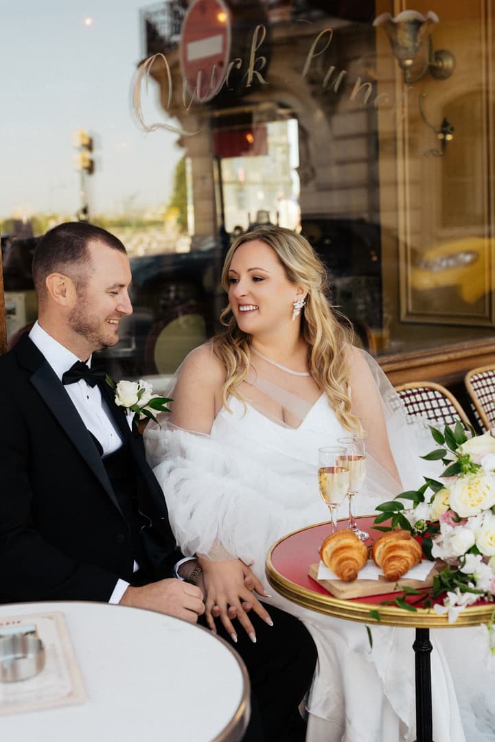 Victoria and Tyler side by side at a red café table with Champagne and croissants at La Frégate