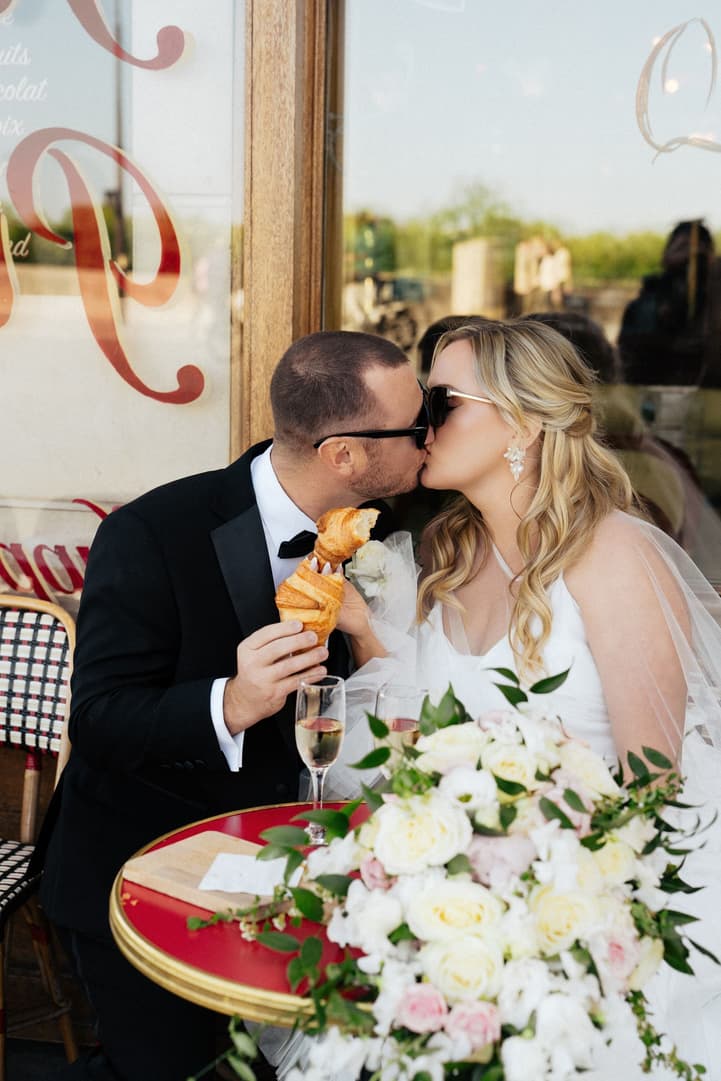 Tyler holds up a croissant and kisses Victoria at their café table at La Frégate